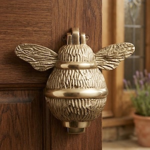 May include: A brass-colored metal door knocker shaped like a bumblebee with textured wings and a rounded body. The knocker is mounted on a dark brown wooden door and features the text CrownWakeMercantile printed in the bottom left corner.