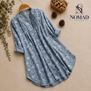 Puede incluir: Una camisa azul de manga larga con botones, estampado floral blanco, cuello mao y pliegues verticales a lo largo de la tapeta frontal. El texto Nomad Styles con un logotipo aparece en la esquina superior derecha de la imagen.