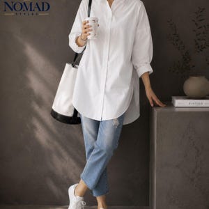 May include: A crisp white long-sleeved button-down shirt with a high-low hemline, paired with light blue distressed denim jeans and white sneakers. A white and black tote bag is carried over the shoulder, and a white cup is held in one hand.