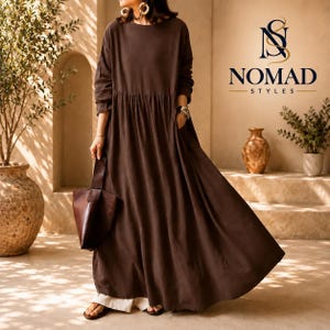 May include: A long-sleeved, floor-length dark brown dress with a gathered waistline and a full, flowing skirt. The dress features a round neckline and is paired with a dark brown leather handbag. The text NOMAD STYLES is visible in the upper right.