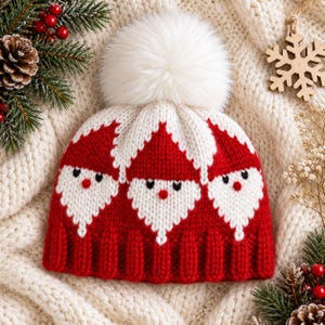 May include: A red and white knit beanie featuring a repeating pattern of Santa Claus faces with white beards, red noses, and black eyes. The hat is topped with a large, fluffy white pom-pom and has a ribbed red knit cuff at the bottom edge.