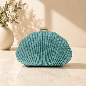 May include: A teal clutch bag shaped like a seashell with a textured, glittery surface and a gold-toned metal clasp at the top. The bag features vertical ridges that mimic the appearance of a shell, providing a distinct, structured, and curved silhouette.