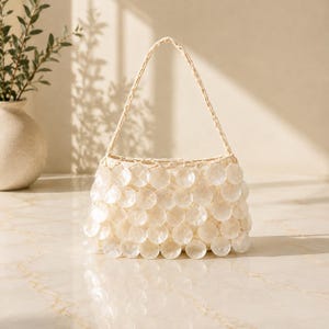 May include: A handbag featuring a structured body covered in overlapping, circular, translucent white shell-like discs. The bag includes a single, braided handle attached to the top, creating a layered, textured appearance with a glossy finish.