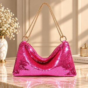 May include: A vibrant hot pink handbag covered in shimmering sequins. The bag features a slouchy, crescent-shaped silhouette with a top zip closure and two gold-toned metal ring attachments connecting to a matching gold-toned chain shoulder strap.