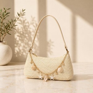May include: A beige woven straw shoulder bag with a single handle and a gold-toned chain draped across the front. The chain is decorated with small white starfish charms and light-coloured seashell accents, featuring a top zip closure.