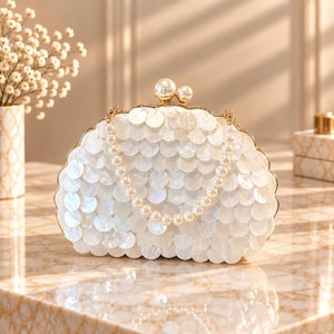 May include: A semi-circular clutch bag covered in overlapping white iridescent shell discs. The bag features a gold-toned metal frame with a dual pearl clasp closure and a detachable handle made of a single strand of white faux pearls.