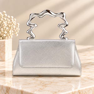 Puede incluir: Un bolso de mano plateado con acabado texturizado y un distintivo asa metálica plateada de forma ondulada. El bolso presenta un cuerpo rectangular estructurado con cierre de solapa, mostrando una estética limpia y una superficie reflectante.