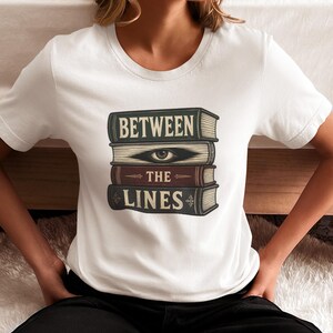 May include: A white short-sleeved t-shirt featuring a graphic of a stack of four books. The top book reads "BETWEEN", the second book displays an eye illustration, the third reads "THE", and the bottom book reads "LINES" in a vintage font.