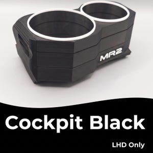 Op de afbeelding: Een zwarte dubbele bekerhouder voor een voertuig, voorzien van twee ronde openingen met witte sierringen. De zijkant van de unit toont de witte tekst MR2. Onder het object staat de tekst Cockpit Black en LHD Only in een wit lettertype.