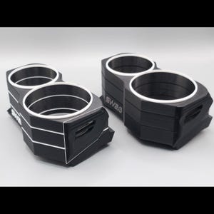 May include: Two black dual cup holders with white circular rims and white accents. Each unit features a geometric, multi-layered design with a side cutout and the text "SWA2" printed in white on the side. The holders are made of a matte material.