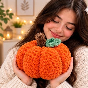 Puede incluir: Una calabaza redonda tejida a ganchillo con hilo grueso y suave. La calabaza presenta una superficie texturizada con costillas verticales, un tallo cilíndrico de color marrón oscuro y una pequeña hoja tejida de color verde claro en la parte superior.