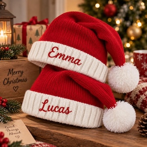 May include: Two red and white knit Santa hats featuring a ribbed cuff and a white pom-pom at the tip. The top hat is embroidered with the name Emma in red script, and the bottom hat is embroidered with the name Lucas in red script.