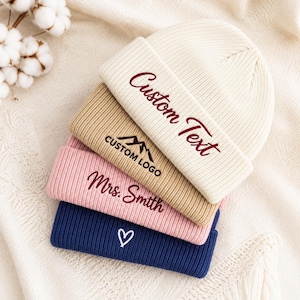May include: A stack of four ribbed knit beanies in cream, tan, light pink, and navy blue. The cream beanie features the text Custom Text, the tan beanie shows a mountain icon with CUSTOM LOGO, the pink beanie says Mrs. Smith, and the navy beanie has a white heart.