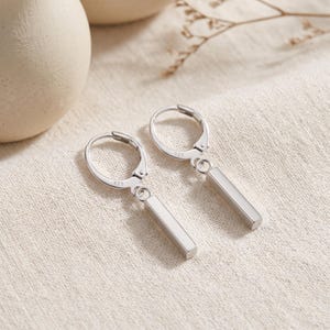May include: A pair of silver hoop earrings featuring a dangling rectangular bar charm. Each earring has a hinged closure and is stamped with the 925 hallmark, indicating sterling silver composition. The design is polished and metallic.