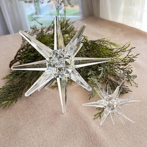May include: Two clear, faceted glass star ornaments with pointed edges. One large star and one smaller star are displayed on a beige surface with green foliage. The stars are decorative and reflect light.