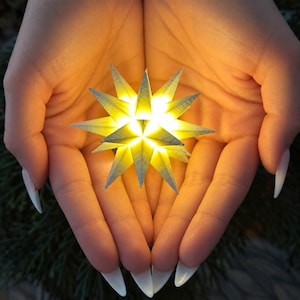 May include: A multi-pointed, three-dimensional star ornament constructed from folded paper strips. The star features a pale, off-white color and emits a warm, bright yellow glow from its center, creating sharp, geometric shadows along its many points.