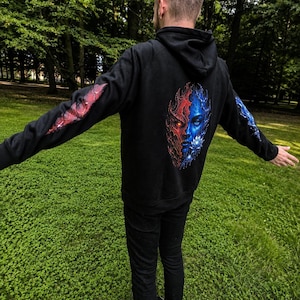 Könnte beinhalten: Ein schwarzer Kapuzenpullover mit einer großen Grafik auf der Rückseite, die ein geteiltes Gesicht in Rot- und Blautönen zeigt. Die rechte Seite ist rot mit einem leuchtenden Auge, die linke Seite ist blau mit einer weißen Lotusblüte. Rote und blaue Akzente befinden sich auf den Ärmeln.