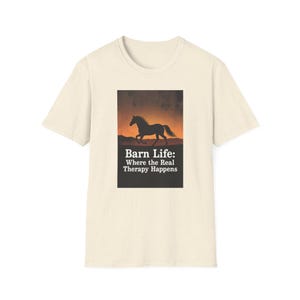 Op de afbeelding: Een crèmekleurig T-shirt met korte mouwen en een rechthoekige grafische print. De afbeelding toont het zwarte silhouet van een paard dat rent tegen een oranje zonsondergang, met de witte tekst: Barn Life: Where the Real Therapy Happens.