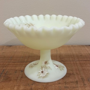 May include: A pale yellow custard glass compote bowl featuring a ruffled, crimped edge and a pedestal base. The exterior of the base and the bowl are decorated with hand-painted white floral motifs with brown centers and green leaves.