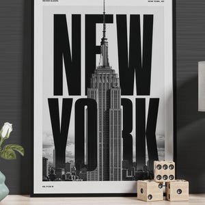 May include: A black and white framed poster featuring the Empire State Building in New York City. The text reads "NEW YORK" in large bold letters, "THE CITY THAT NEVER SLEEPS", "EMPIRE STATE NEW YORK, NY", and the coordinates "40.7128 N".