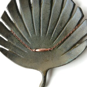 May include: A handmade, leaf-shaped ceramic dish in shades of green and gray, holding a delicate copper chain necklace with a cylindrical pendant. The dish has a textured surface and is set against a white background.