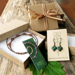 May include: Jewelry set in gift boxes. The set includes a necklace with brown beads and copper accents, leaf-shaped earrings, and a copper wire bracelet with green beads. The boxes are brown and tied with raffia.