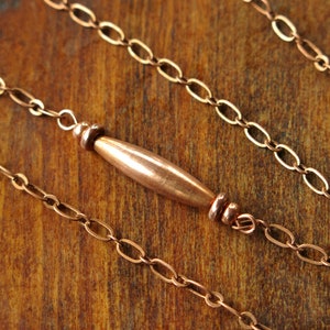 May include: Close-up of a copper necklace with a central barrel-shaped bead and delicate chain links. The warm, reddish-brown metal contrasts against a wooden background, highlighting the jewelry's design.