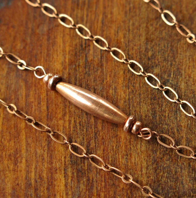 May include: Close-up of a copper necklace with a central barrel-shaped bead and delicate chain links. The warm, reddish-brown metal contrasts against a wooden background, highlighting the jewelry's design.