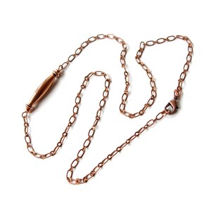 May include: A copper-colored necklace with an oval link chain and a cylindrical pendant. The necklace has a lobster clasp closure. The pendant is a smooth, elongated shape with decorative details at each end. The jewelry is displayed on a white background.