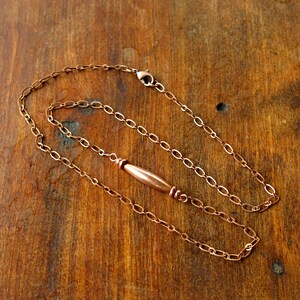 May include: A copper-colored necklace with an oval chain and a central, elongated bead. The necklace is displayed on a wooden surface, showcasing its warm, metallic tones. The clasp is visible, indicating the necklace's design.