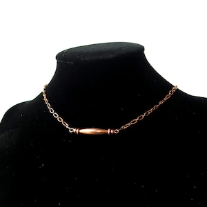 May include: A copper-colored necklace with a bar pendant. The necklace is displayed on a black mannequin. The chain is made of small, oval links. The bar pendant is cylindrical with a slightly darker hue.