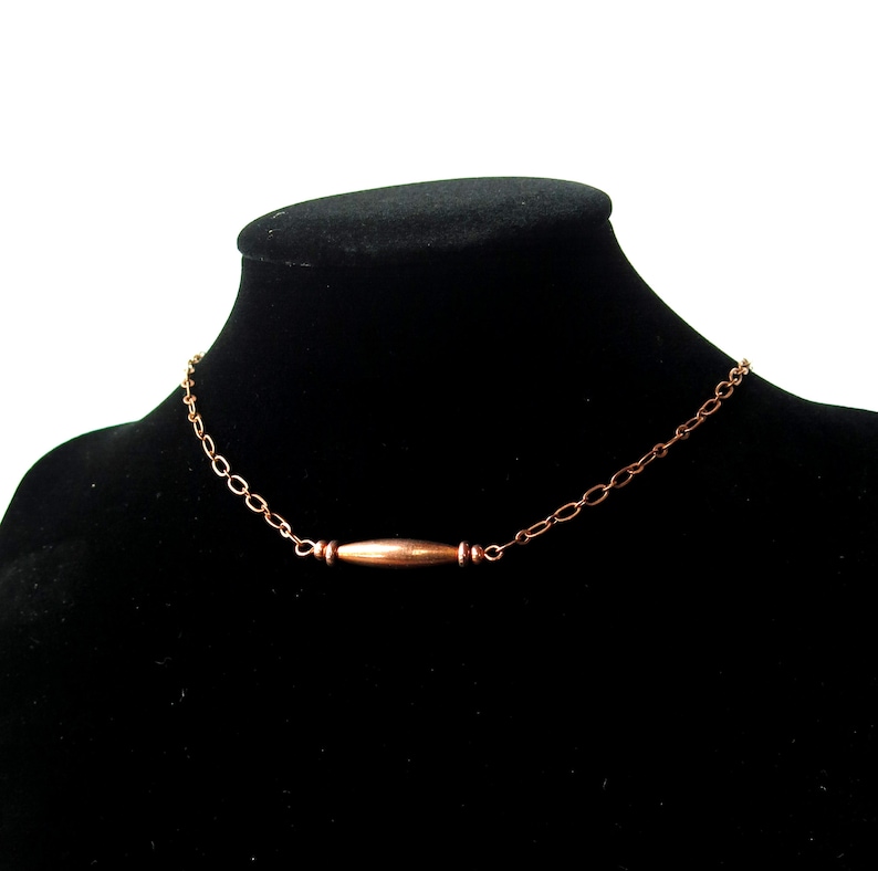 May include: A copper-colored necklace with a bar pendant. The necklace is displayed on a black mannequin. The chain is made of small, oval links. The bar pendant is cylindrical with a slightly darker hue.
