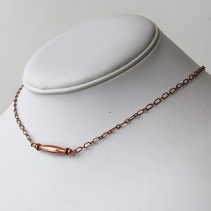 May include: A copper-colored necklace with a central, elongated bead and delicate chain links. The necklace is displayed on a white mannequin bust, showcasing its design and craftsmanship. The jewelry piece is simple and elegant.