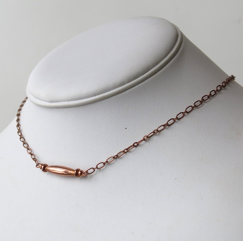 May include: A copper-colored necklace with a central, elongated bead and delicate chain links. The necklace is displayed on a white mannequin bust, showcasing its design and craftsmanship. The jewelry piece is simple and elegant.