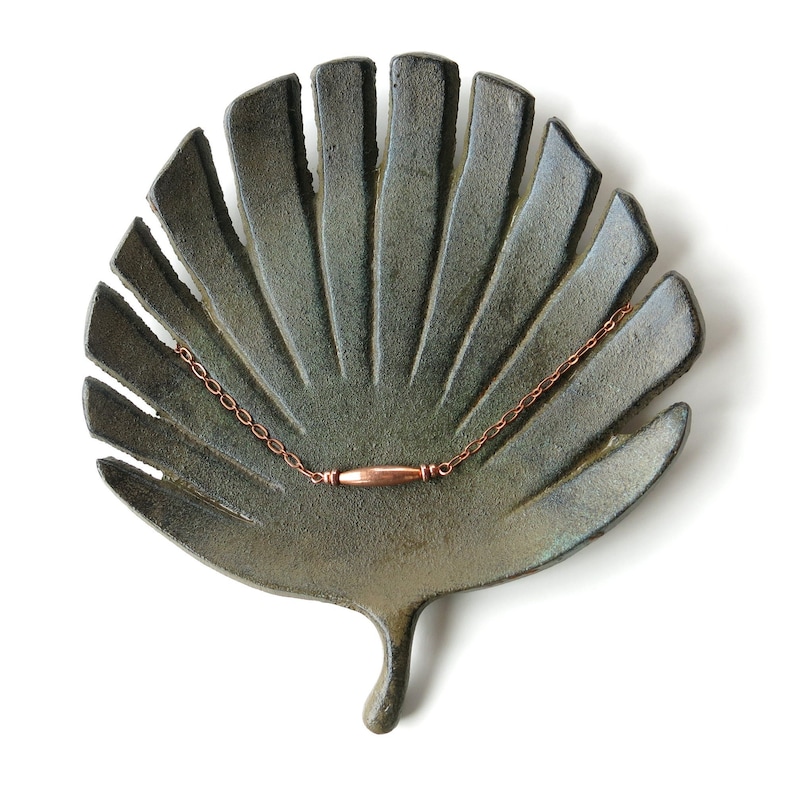 May include: A handmade, leaf-shaped ceramic dish in shades of green and gray, holding a delicate copper chain necklace with a cylindrical pendant. The dish has a textured surface and is set against a white background.
