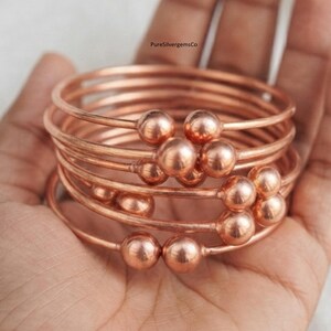Op de afbeelding: Een stapel van meerdere koperkleurige metalen armbanden, elk met een gladde, gepolijste afwerking en twee bolvormige uiteinden. De tekst PureSilvergemsCo is zichtbaar in het midden bovenaan de afbeelding boven de metalen armbanden.
