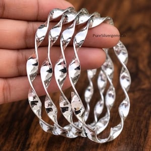 May include: A set of four silver-colored bangles featuring a continuous twisted, spiral design with a polished, reflective surface. The metal bracelets have a uniform, undulating shape throughout their circumference, displaying the text PureSilvergemsCo.