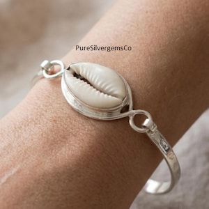 Può includere: Un bracciale in argento con una conchiglia di ciprea bianca centrale incastonata in una cornice d'argento lucido. Il cinturino è realizzato in argento liscio e si collega all'incastonatura della conchiglia con maglie circolari. Il testo PureSilvergemsCo è visibile.