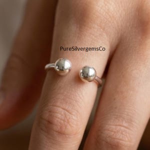 Op de afbeelding: Een zilveren open ring met twee gepolijste bolvormige uiteinden. De band is glad en metaalachtig, met de tekst PureSilvergemsCo zichtbaar boven de ring. Het ontwerp is eenvoudig en cirkelvormig, met een reflecterend oppervlak op de bollen.