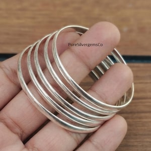 Peut inclure: Un ensemble de cinq bracelets joncs en argent lisse et poli empilés ensemble. Les anneaux métalliques présentent un profil arrondi et une finition métallique réfléchissante. Le texte PureSilvergemsCo est visible sur le bracelet supérieur.