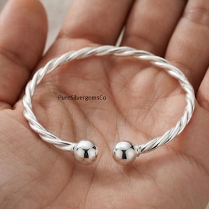 Può includere: Un bracciale a fascia in argento con un design a corda intrecciata e due estremità sferiche lucide. La superficie metallica è riflettente e liscia. Il testo PureSilvergemsCo è stampato in nero sulla curva interna del bracciale in argento.
