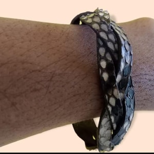 Könnte beinhalten: Ein geflochtenes Armband aus einem strukturierten Material mit Schlangenhautmuster in Schwarz, Weiß und Grau. Das Design besteht aus mehreren ineinander verschlungenen Strängen, die ein durchgehendes, flexibles Band mit einer schuppenartigen Optik bilden.