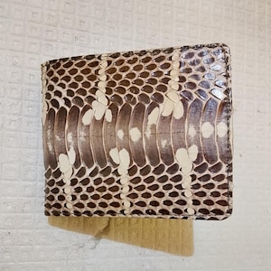 Op de afbeelding: Een bi-fold portemonnee met een gestructureerd slangenleerpatroon in tinten bruin, crème en donkergrijs. De buitenkant toont een herhalend schubbenontwerp met een centrale verticale strook van grotere, lichtere schubben over het oppervlak.