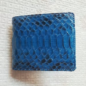May include: A bifold wallet featuring a vibrant blue snakeskin texture with a distinct scale pattern. The wallet has a rectangular shape with stitched edges, showcasing a deep cobalt blue colour with darker black accents between the individual scales.