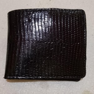 Op de afbeelding: Een donkerbruine bifold portemonnee met een gestructureerd, reliëfoppervlak dat het uiterlijk van hagedissenhuid-schubben nabootst. De portemonnee heeft een rechthoekige vorm met afgeronde hoeken en een glanzende afwerking die het licht reflecteert.