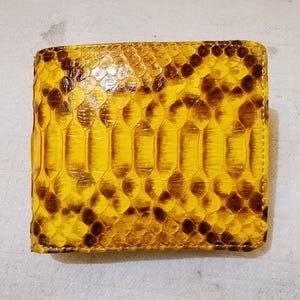 May include: A square bifold wallet featuring a vibrant yellow and dark brown snakeskin pattern. The exterior displays a repeating scale texture across the entire surface, with stitched edges defining the rectangular shape of the accessory.