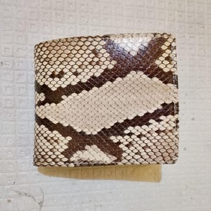 Könnte beinhalten: Ein quadratisches Portemonnaie mit zwei Fächern und einem strukturierten Schlangenledermuster in Creme-, Beige- und Dunkelbrauntönen. Die Oberfläche zeigt ein sich wiederholendes, rautenförmiges Schuppenmuster über die gesamte Außenseite.