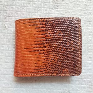 Op de afbeelding: Een bi-fold portemonnee met een getextureerd oppervlak en een kleurverloop dat overgaat van lichtbruin aan de linkerkant naar diep donkerbruin aan de rechterkant. De buitenkant vertoont een opvallend patroon van kleine, reptielachtige schubben.