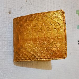 May include: A bi-fold wallet featuring a metallic gold finish and a distinct, raised scale-like texture across the entire exterior surface. The wallet has a rectangular shape with stitched edges and a consistent, reflective golden colour throughout.