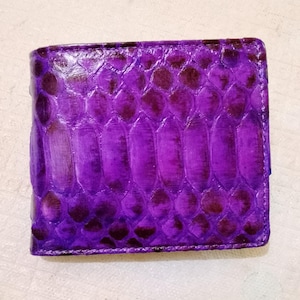 May include: A bi-fold wallet featuring a vibrant purple and dark violet snakeskin pattern. The exterior displays a distinct, repeating scale texture across the entire surface, with a glossy finish that highlights the deep colour variations.